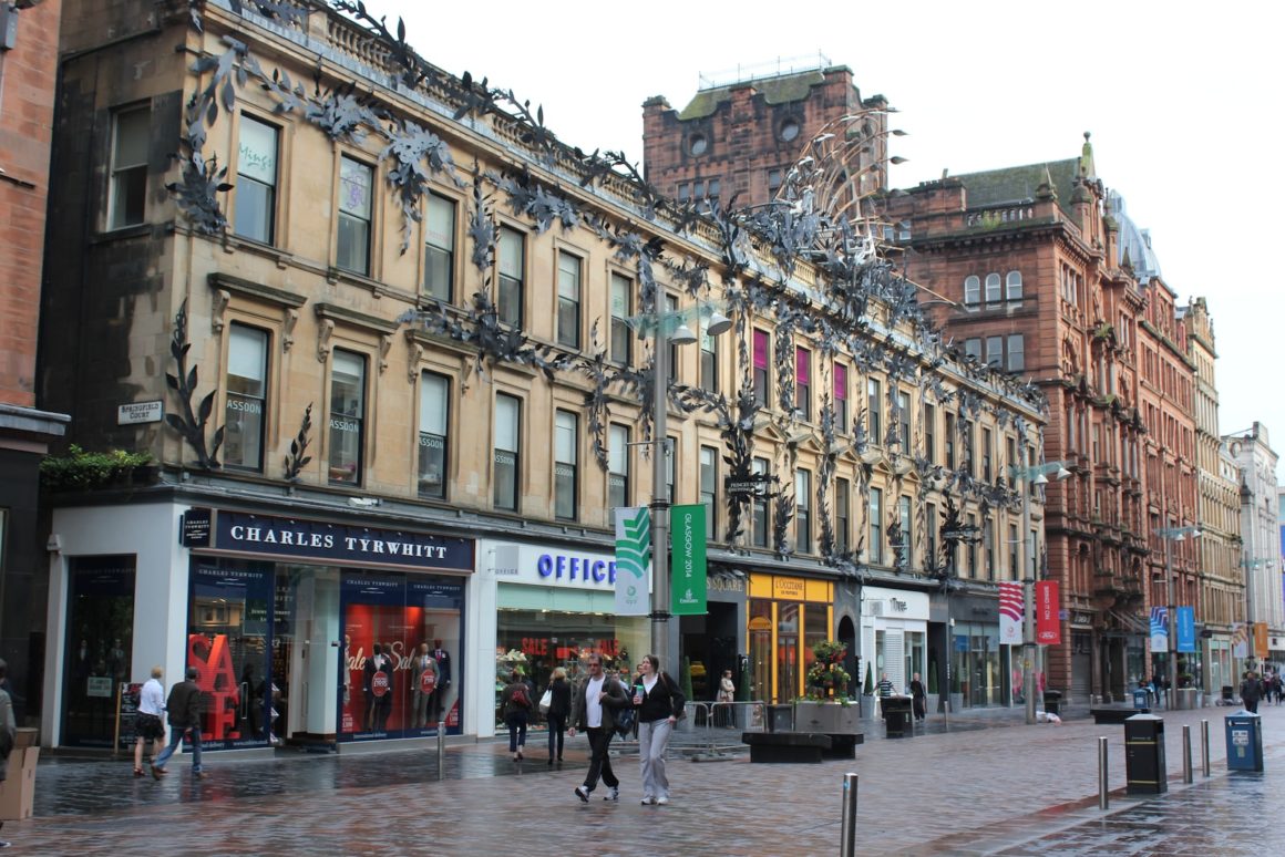 Bruisende scène op Princes Square, Glasgow, met mensen die langs de elegante winkelstraat met chique boetieks en historische architectuur slenteren, die het bruisende stadsleven vastlegt.