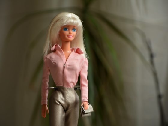 Barbie doll wearing a professional pink business suit, complete with a fitted blazer and skirt. She accessorizes the outfit with a matching pink purse, stiletto heels, and stylish sunglasses. Barbie's blond hair is pulled back into a neat ponytail, exuding confidence and elegance in the office setting.