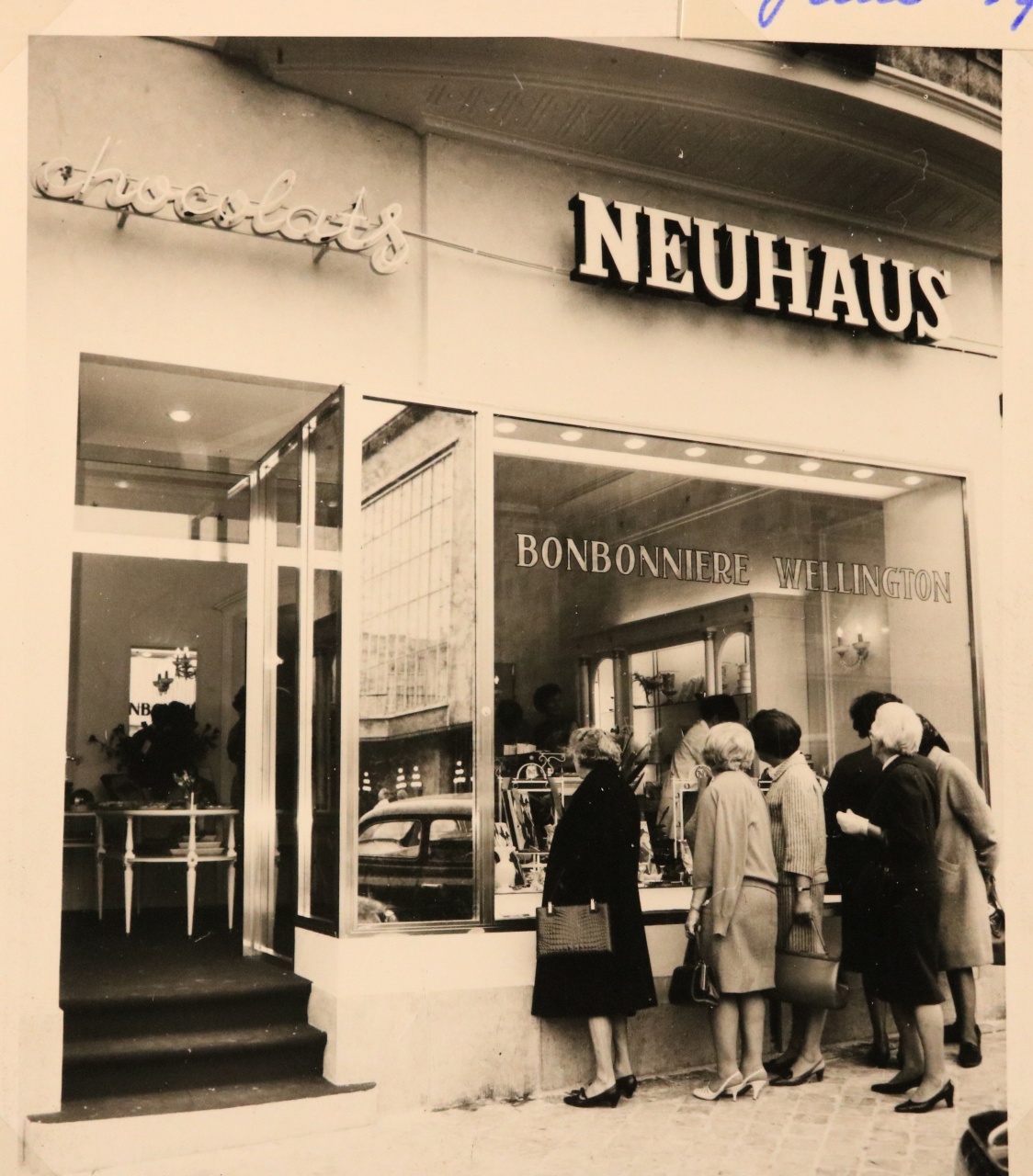 Historic black-and-white photo of the exterior of the Neuhaus chocolate shop, with customers standing in front of the window