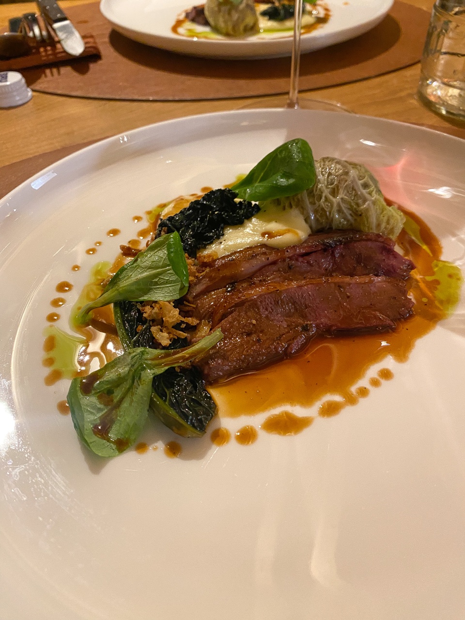 A tasty-looking dish served on a white plate. It appears to be a piece of roasted meat, possibly duck, garnished with a creamy sauce and crispy toppings. On the side are added green leaves and cooked vegetables, probably spinach or a similar leafy vegetable. The whole thing is complemented by a dainty gravy or sauce artistically drizzled around the meat. The dish exudes craftsmanship and sophistication, typical of a high-end restaurant.