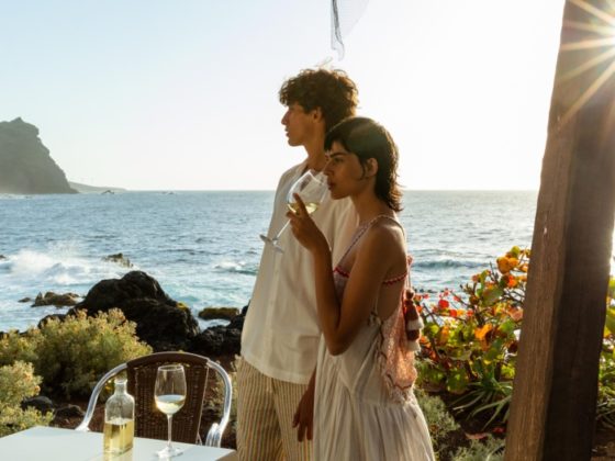 Een man en een vrouw genieten van een glas wijn bij een tafel aan de kust van Tenerife, met uitzicht op de oceaan en rotsachtige kliffen in de achtergrond. De zon schijnt helder, waardoor een serene en romantische sfeer ontstaat.
