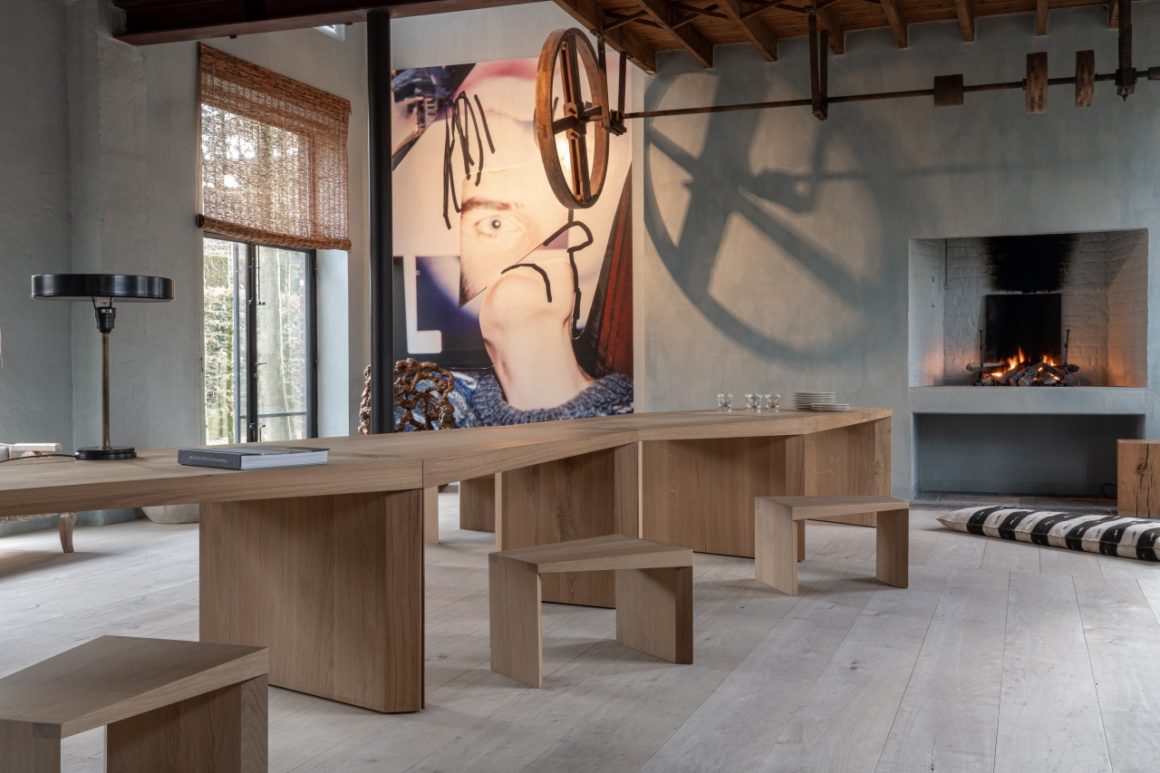 A warm and sculptural interior featuring The Endless Table by Bernard De Clerck for Collected by Obumex, crafted in natural oak with a zigzag modular design, styled with minimalist wooden stools, a black table lamp, and a modern fireplace adding cozy elegance. A large contemporary artwork and exposed ceiling beams complete the artistic ambiance.