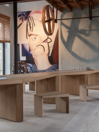 A warm and sculptural interior featuring The Endless Table by Bernard De Clerck for Collected by Obumex, crafted in natural oak with a zigzag modular design, styled with minimalist wooden stools, a black table lamp, and a modern fireplace adding cozy elegance. A large contemporary artwork and exposed ceiling beams complete the artistic ambiance.