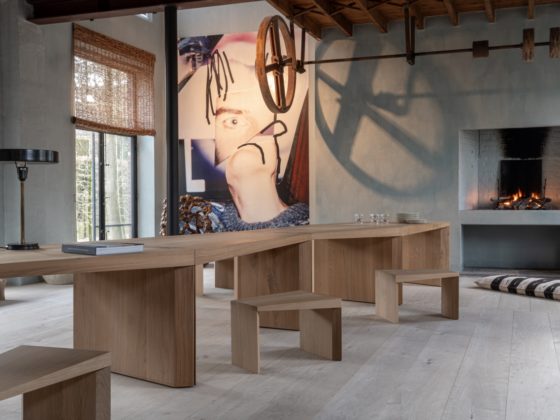 A warm and sculptural interior featuring The Endless Table by Bernard De Clerck for Collected by Obumex, crafted in natural oak with a zigzag modular design, styled with minimalist wooden stools, a black table lamp, and a modern fireplace adding cozy elegance. A large contemporary artwork and exposed ceiling beams complete the artistic ambiance.
