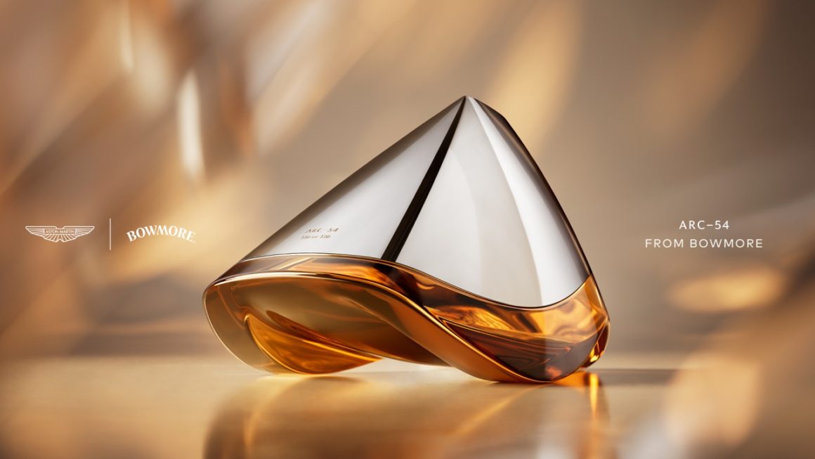 Close-up of the futuristic Bowmore ARC-54 whisky decanter, designed in collaboration with Aston Martin. The hand-blown vessel features a sleek silver top and amber-colored whisky below, set against a warm, golden-toned background. On the left are the Aston Martin and Bowmore logos; on the right, the text reads “ARC-54 from Bowmore