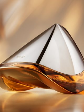 Close-up of the futuristic Bowmore ARC-54 whisky decanter, designed in collaboration with Aston Martin. The hand-blown vessel features a sleek silver top and amber-colored whisky below, set against a warm, golden-toned background. On the left are the Aston Martin and Bowmore logos; on the right, the text reads “ARC-54 from Bowmore