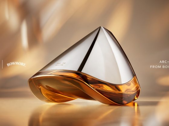 Close-up of the futuristic Bowmore ARC-54 whisky decanter, designed in collaboration with Aston Martin. The hand-blown vessel features a sleek silver top and amber-colored whisky below, set against a warm, golden-toned background. On the left are the Aston Martin and Bowmore logos; on the right, the text reads “ARC-54 from Bowmore
