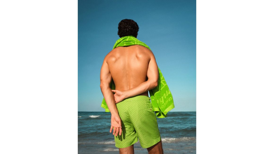 Back view of a man in green patterned swim shorts holding a matching towel, standing on the beach facing the ocean under a clear blue sky.