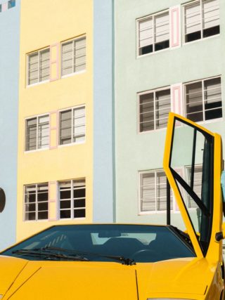Man in turquoise summer outfit leaning on yellow Lamborghini Countach with door open, talking on phone near colorful Miami Beach architecture.