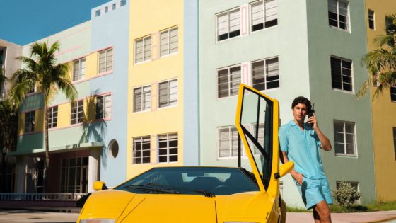 Man in turquoise summer outfit leaning on yellow Lamborghini Countach with door open, talking on phone near colorful Miami Beach architecture.