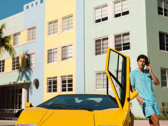 Man in turquoise summer outfit leaning on yellow Lamborghini Countach with door open, talking on phone near colorful Miami Beach architecture.