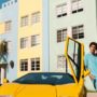 Man in turquoise summer outfit leaning on yellow Lamborghini Countach with door open, talking on phone near colorful Miami Beach architecture.