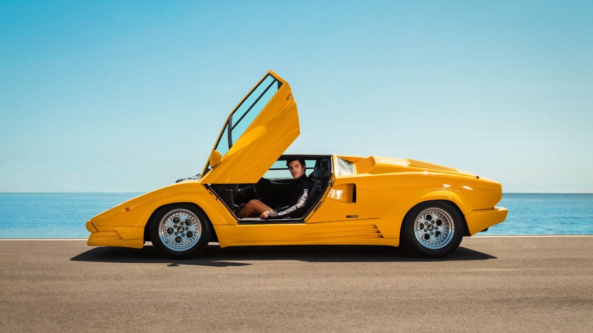 Man sitting inside a yellow Lamborghini Countach with the scissor door open, parked by the sea under bright Miami sunlight.