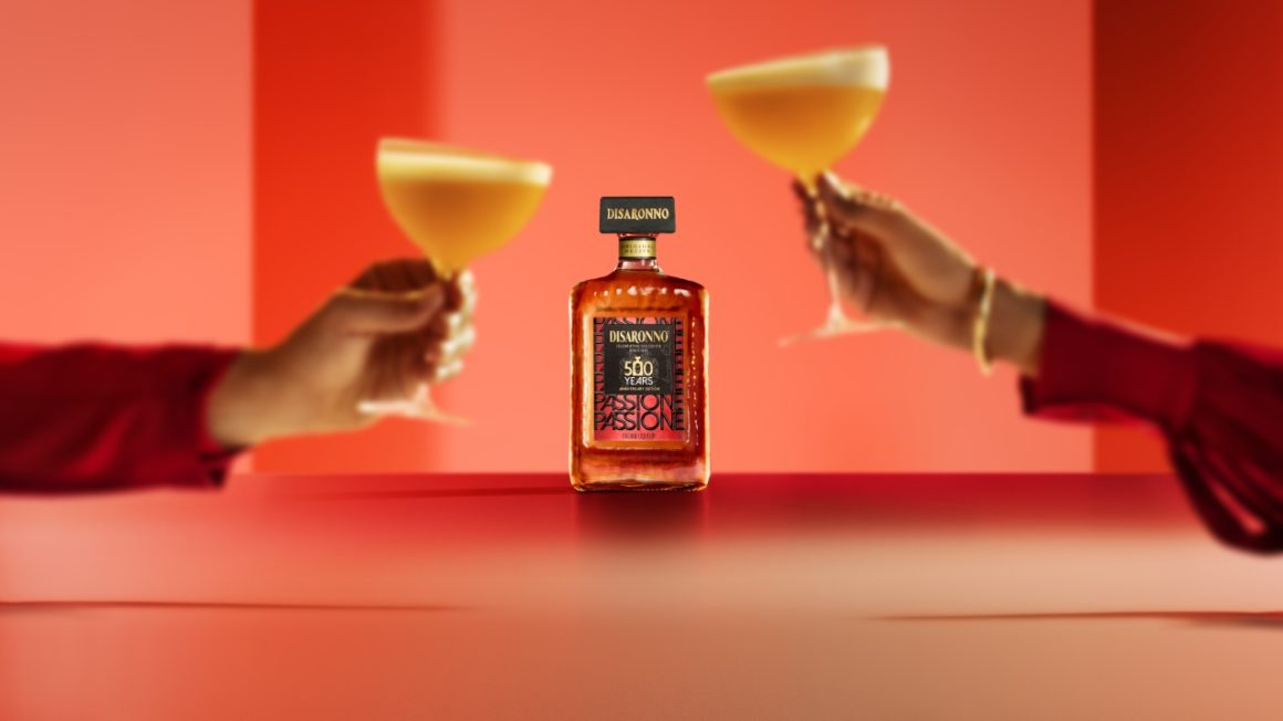 Disaronno Passione limited edition bottle in the center, with two hands raising elegant cocktails for a toast, set in a warm red-toned atmosphere.