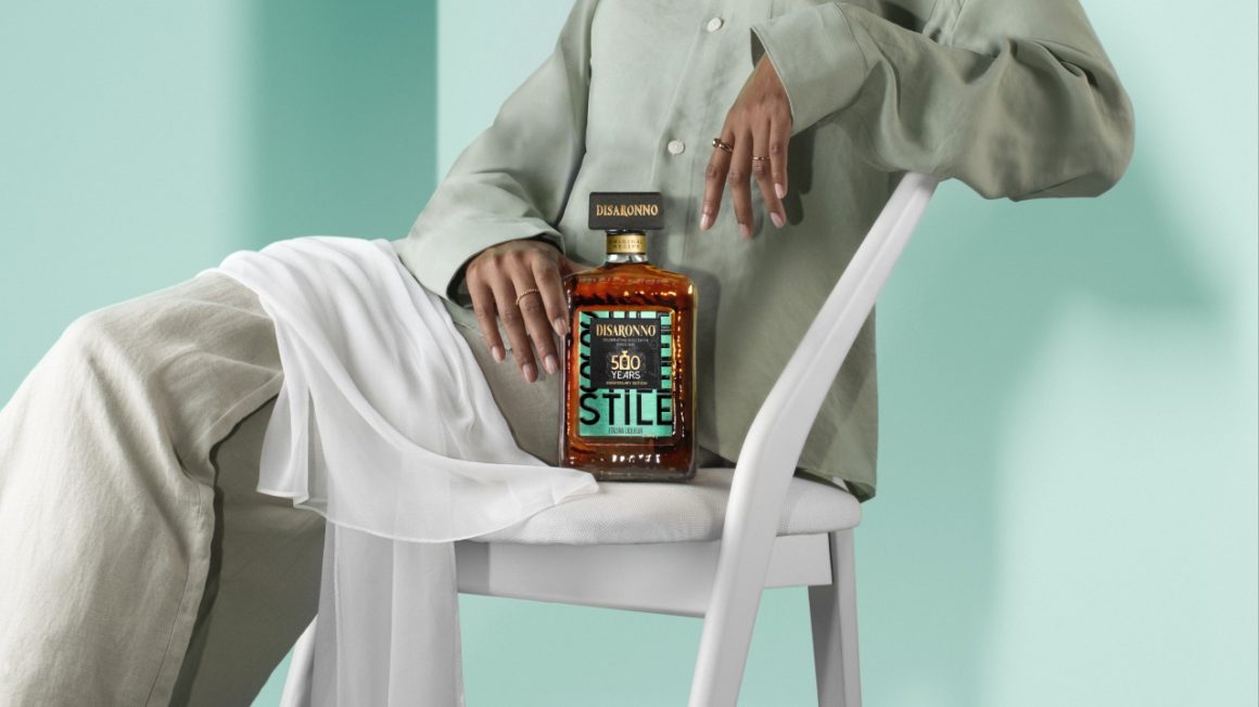 Disaronno Stile limited edition bottle resting on the lap of a woman in a minimalist mint green outfit, seated on a white chair with a flowing white scarf.